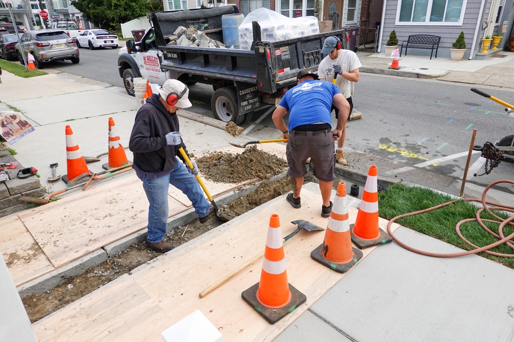 Crew replacing a residential driveway with excavation work, safety cones, and construction equipment on a city street.